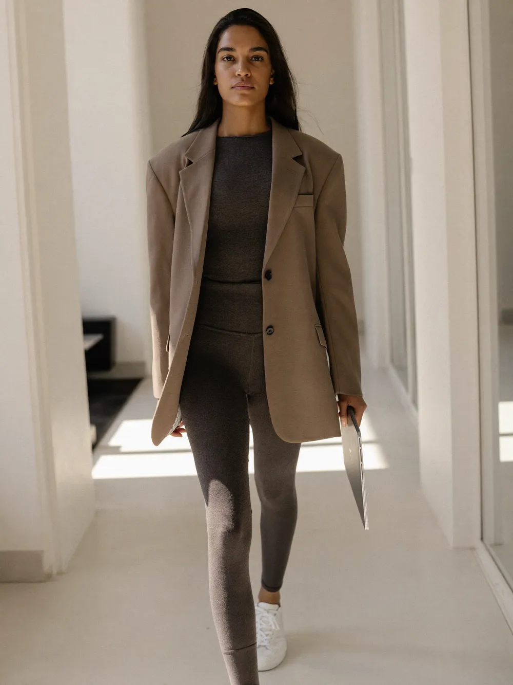 Mujer con blazer hueso sobre leggings carbón caminando por un pasillo luminoso con una laptop